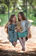 Happy laughing kids girls sisters with long hair enjoying a swing ride with a teddy bear toy on a sunny summer day