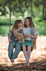 Happy laughing kids girls sisters with long hair enjoying a swing ride with a teddy bear toy on a sunny summer day