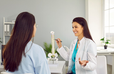 Friendly female physiotherapist in hospital office advises young woman on intervertebral hernia. Smiling woman in white medical coat shows anatomical model of spine to her female patient.