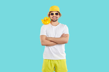 Funny cheerful guy enjoying his summer holiday. Studio portrait of happy young man in white T shirt, yellow shorts, bucket hat and sun glasses standing with toy rubber duck on his shoulder and smiling