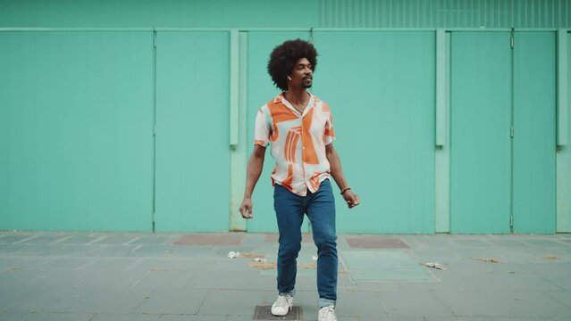 Cheerful young African American man wearing shirt listening to music in wireless headphones and dancing on light blue wall background. Slow motion, camera zooming
