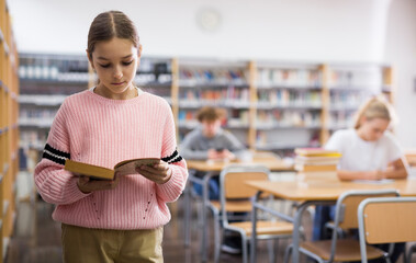 Portrait of a ten-year-old schoolgirl standing near a bookcase in the school library with a book in her hands