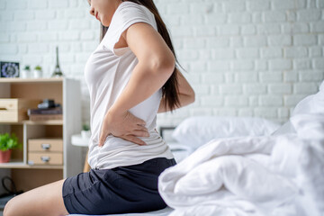 Asian woman sitting in bed touching her back, feeling back pain in the morning. She suffered from...