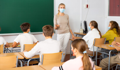 Teacher in face mask explaining subject to pupils during lesson in classroom.