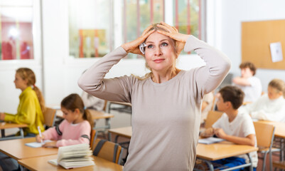 Waist-up portrait of irritated female teacher standing in classroom and holding head with hands.