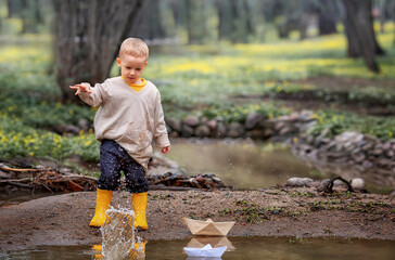 Portrait of adorable little kid boy in rubber boots launch paper boats in a puddle in spring. Origami.