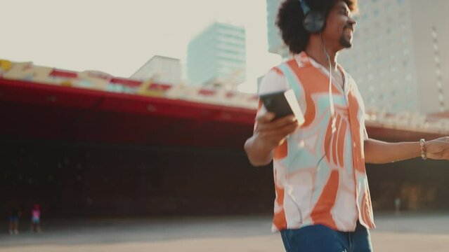 Close-up of cheerful young African American man wearing shirt listening to music in headphones and dancing on urban city background. Lifestyle concept. Backlight Slow motion