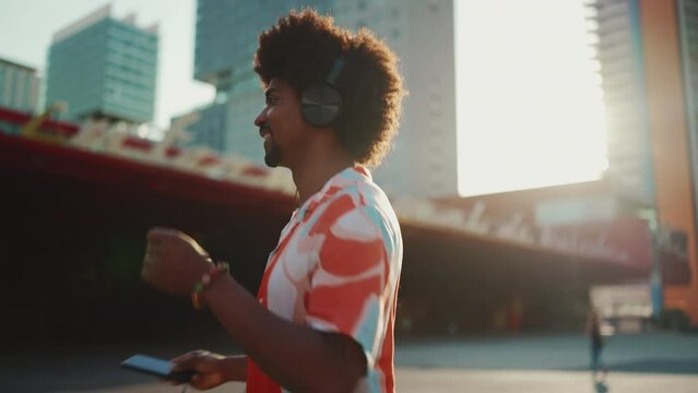 Close-up of cheerful young African American man wearing shirt listening to music in headphones and dancing on urban city background. Lifestyle concept. Backlight Slow motion