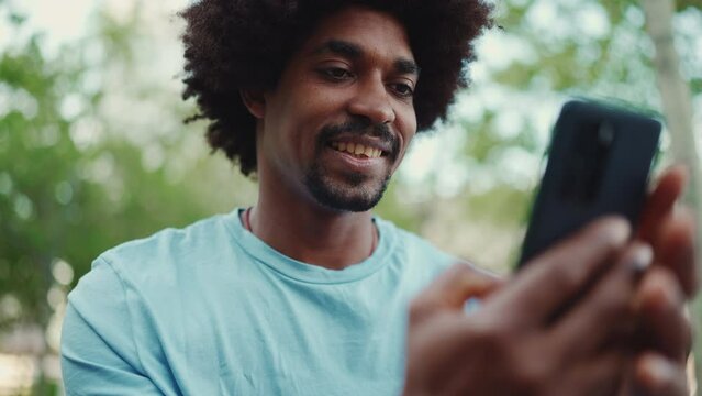 Closeup portrait of young African American man in light blue t-shirt sitting on city park bench and using his smartphone. Man looks at photos videos in his mobile phone. lifestyle concept. Slow motion