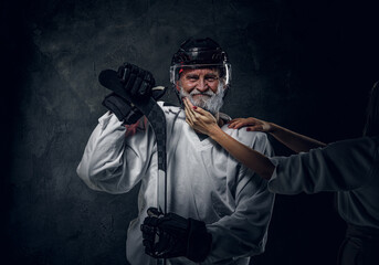 Shot of happy old man hockey player with hockey stick being touched by woman against dark...