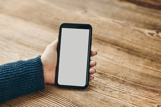 Female Hand Holding Smartphone Mockup With Solid White Color Blank Empty Isolated Screen On Wooden Background. Person Using Technology. Mobile Phone With Copy Space Left