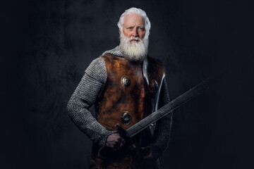 Studio shot of medieval elderly warrior dressed in armored clothes holding long sword.