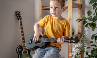 Cute cheerful boy is playing ukulele guitar in the music room. Joyful learning to play musical...