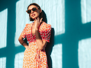 Young beautiful confident female in trendy summer red dress. Sexy carefree woman posing near blue wall in studio. Positive model having fun. Cheerful and happy. At sunny day. Shadow from window