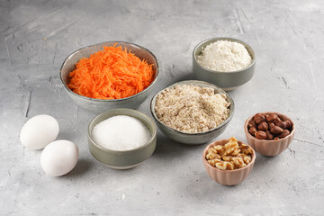 Ingredients for baking a carrot cake: finely grated carrots, grounded hazelnuts and almonds, chopped walnuts, white flour, eggs, sugar on grey background