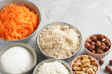 Ingredients for baking a carrot cake: finely grated carrots, grounded hazelnuts and almonds, chopped walnuts, white flour, eggs, sugar on grey background