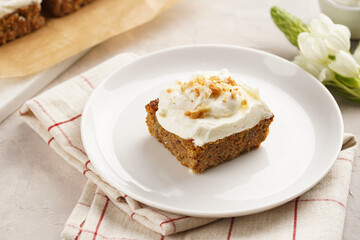 Homemade pastry carrot-walnut cake with grounded almonds and hazelnuts and white cream cheese top layer on white plate on pink checkered napkin