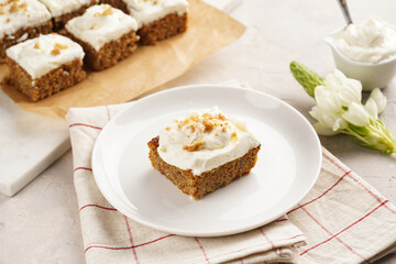 Homemade pastry carrot-walnut cake with grounded almonds and hazelnuts and white cream cheese top layer on white plate on pink checkered napkin