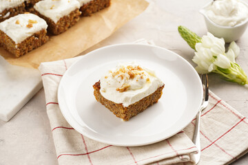Homemade pastry carrot-walnut cake with grounded almonds and hazelnuts and white cream cheese top layer on white plate on pink checkered napkin