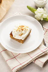 Homemade pastry carrot-walnut cake with grounded almonds and hazelnuts and white cream cheese top layer on white plate on pink checkered napkin