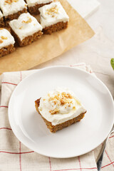 Homemade pastry carrot-walnut cake with grounded almonds and hazelnuts and white cream cheese top layer on white plate on pink checkered napkin