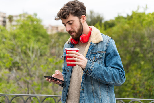 Young man watching smartphone with bicchiere di plastica in mano and wearing headset – guy in casual outfit with wireless headset – happy male using smartphone in the park 