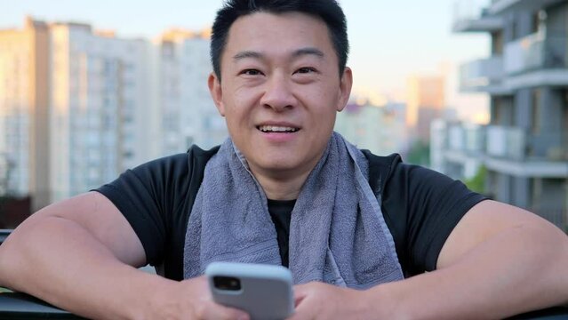 Handsome asian man with a towel around his neck holds a mobile phone looking to camera. Satisfied Asian man smiles after workout resting after a strength training. The concept of sports.