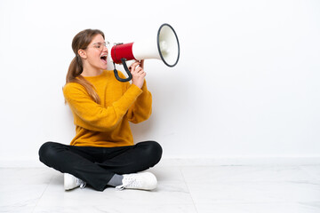 Young caucasian woman sitting on the floor isolated on white background shouting through a megaphone