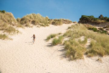 A woman wearing a swimming suit and a hat and her daughter run down  a white sand beach under a blue  in Camusdarach Beach, Mallaig, Scottish Highlands, UK