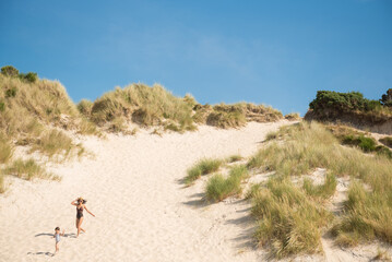 A young woman wearing a swimming suit and a hat and her daughter's run down  a white sand beach under a blue  in Camusdarach Beach, Mallaig, Scottish Highlands, UK