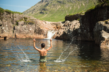 A woman wearing a green swimming suit splashing water with both hands while bathing in the River Etive in Glen Etive, Scottish Highlands, UK, with a waterfall and a mountain in the background.