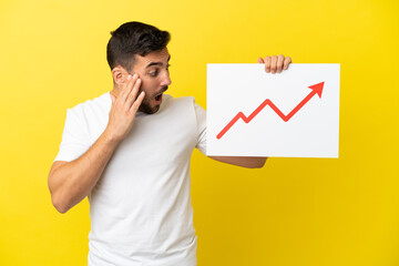 Young handsome caucasian man isolated on yellow background holding a sign with a growing statistics arrow symbol with surprised expression