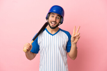 Baseball player with helmet and bat isolated on pink background smiling and showing victory sign