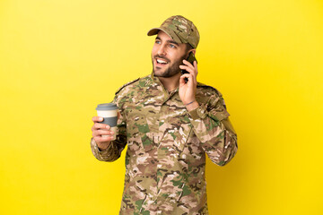 Military man isolated on yellow background holding coffee to take away and a mobile