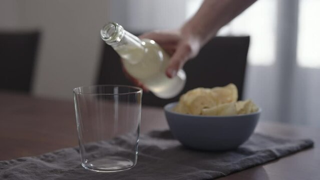 Slow motion man pour ginger beer into tumbler glass with blue bowl full of potato chips