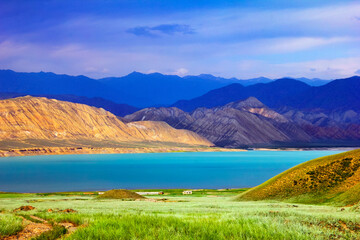 Beautiful scenic view with green field and colorful blue water of Toktogul lake under barren yellow and violet mountains at the background of cloudy sky, Tien Shan range, Kyrgyzstan, Central Asia