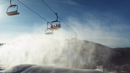 Using ski lift, snow cannon, bottom view on sunny morning. Artificial snow sprayed in sun's rays, of bright blue sky. Concept of winter recreation at ski resort. Chair lift skiers head to slope