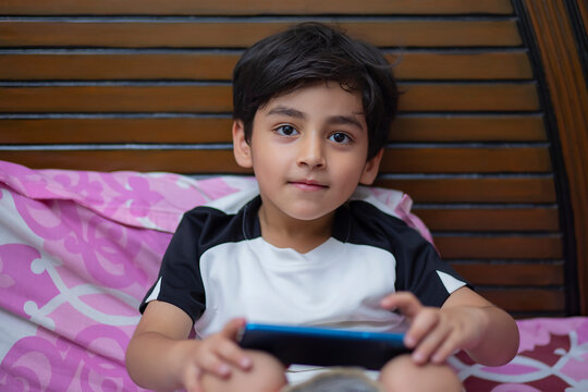 Close-up portrait of little boy looking at camera while lying on bed with smartphone