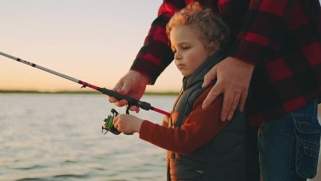 curly little boy is learning to fishing by rod, careful father or grandpa is helping, happy childhood moments