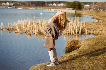 Happy beautiful blonde woman with open arms standing near river bank alone on cool sunny day,...
