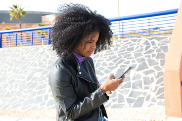Young, beautiful Afro-American woman with mobile phone shopping online, checking her email and social networks in the park while having a coffee. The woman is wearing casual and comfortable clothes.