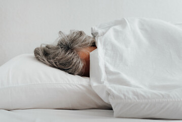 Close-up of frozen caucasian woman with gray hair wrapped up in blanket lying on bed in bedroom. Low temperature, uncomfortable concept
