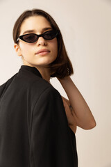 Portrait of young stylish woman in black jacket and trendy sunglasses posing isolated over grey studio background.