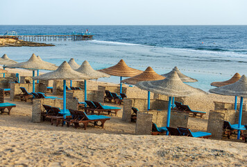 Sandy beach of the Red Sea with straw umbrellas and deck chairs near Marsa Alam, Egypt, Africa. Seascape and waves breaking on the coral reef.