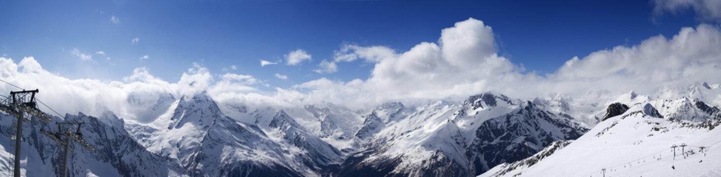 Panoramic view on ski slope and cloudy mountains at sun day