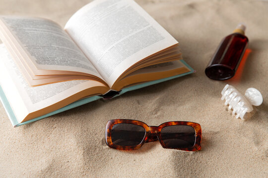 leisure and summer holidays concept - open book, sunscreen, hair clip and sunglasses on beach sand