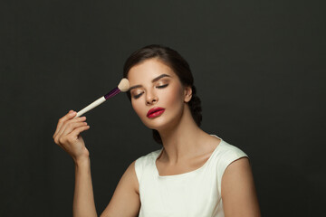 Obraz premium Beautiful glamorous gorgeous elegant young woman with perfect skin applying makeup with a brush against a black background
