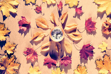 Time change in Autumn. Wooden alarm clock with dry Fall leaves on recycled cardboard. Top view, flat lay, natural sunlight with shadows.