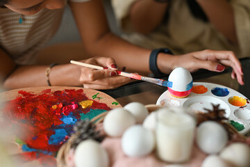 Close up with Asian female painting duck's egg on color palette over blurred Easter holiday decoration background.