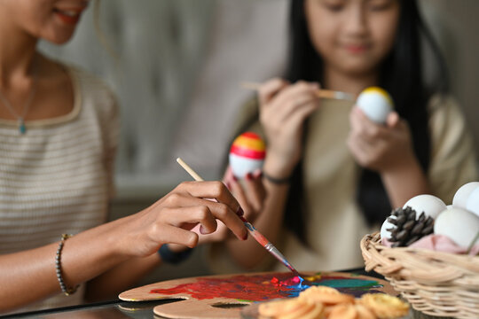 Close up with Asian female hands holding Easter egg and paintbrush while painting duck's egg with over blurred family member background.